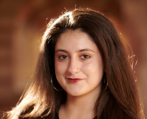 A woman with dark brown hair looks directly at the camera. She is wearing dark pink lipstick, silver hoop earrings and a black button down.